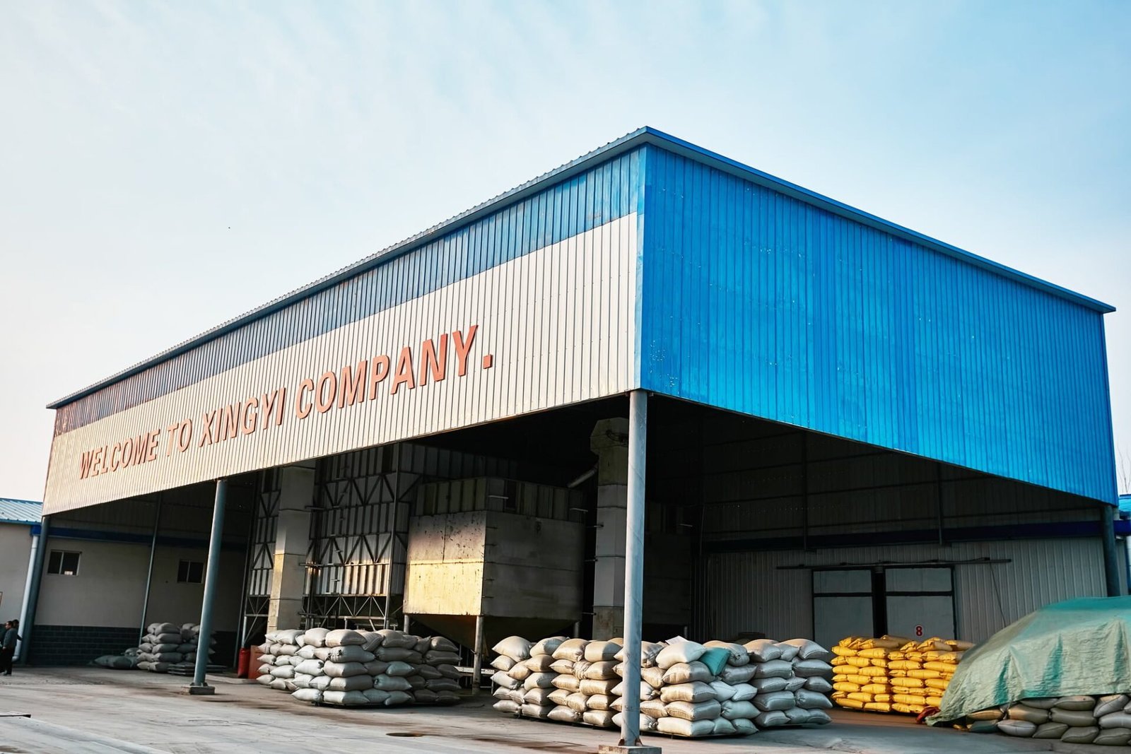 Exterior view of a large blue and white industrial building with 'WELCOME TO XINGYI COMPANY' written on its facade. Stacks of large sacks, likely containing agricultural products, are visible in front of the building. The structure appears to be a warehouse or processing facility for agricultural goods.