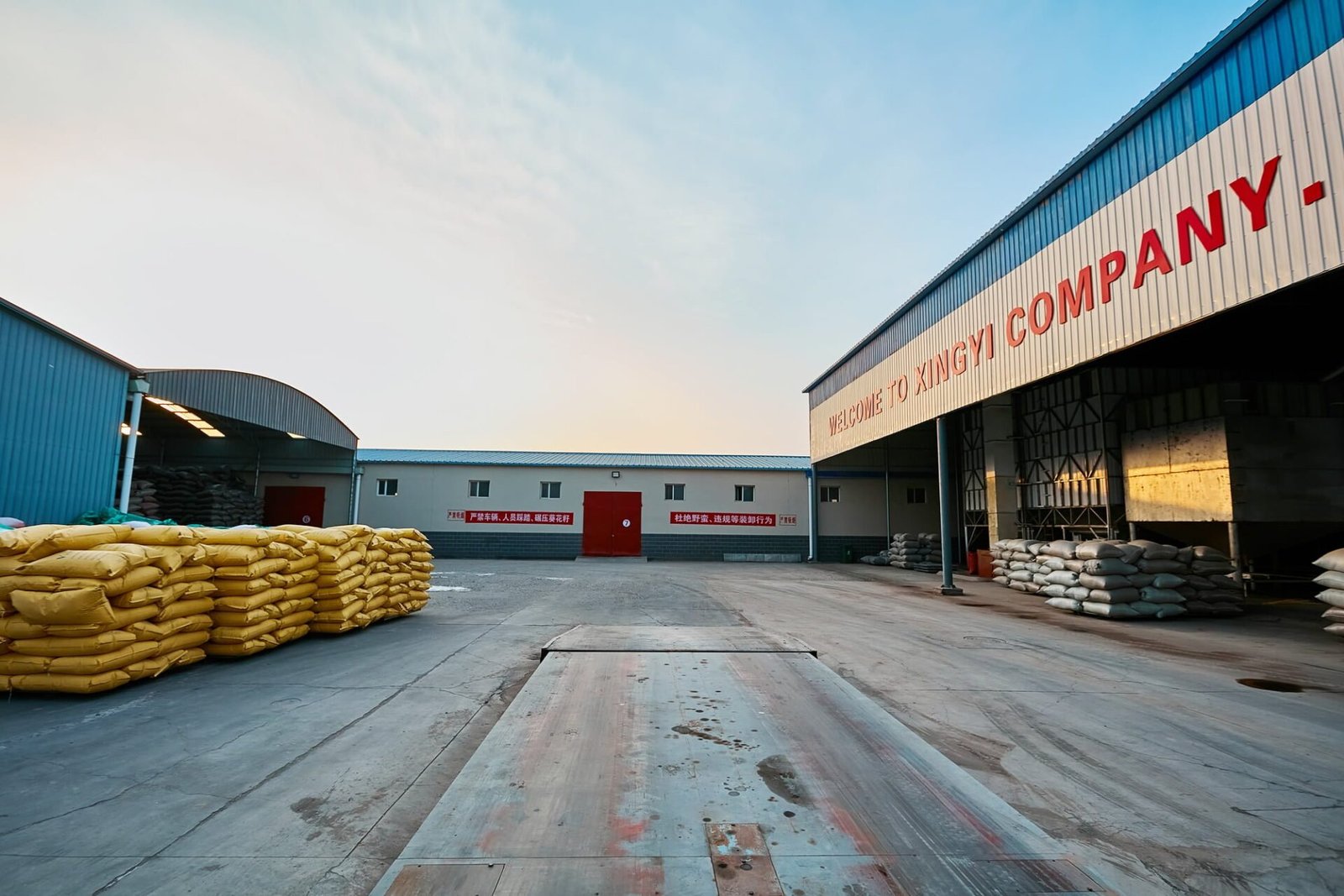 Exterior view of XingYi Company's industrial facility. Large warehouse buildings with blue and white exteriors are visible, with 'WELCOME TO XINGYI COMPANY' in red letters on one building. Stacks of yellow and white sacks, likely containing agricultural products, are seen in the foreground. The image shows a spacious courtyard area with Chinese text visible on a central building.