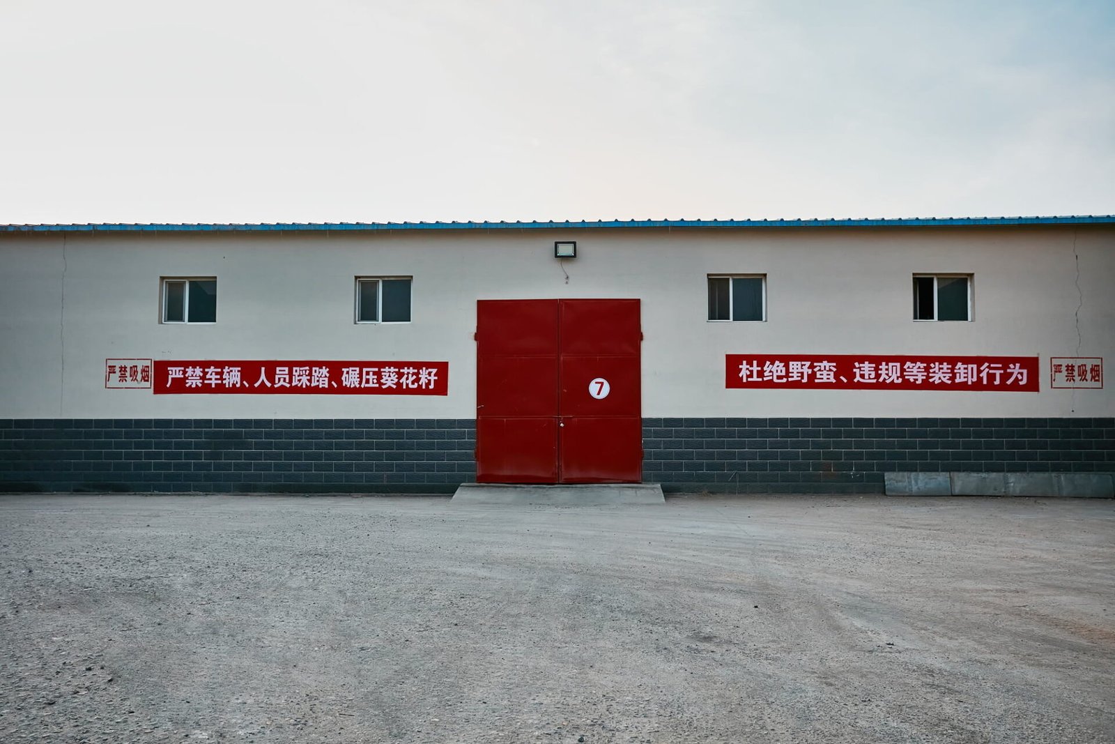 Front view of a white industrial building with a blue roof. It features a central red door marked with the number 7, four windows, and red Chinese text on either side of the door. The text appears to be safety regulations. The building is set against a clear sky with an empty foreground.