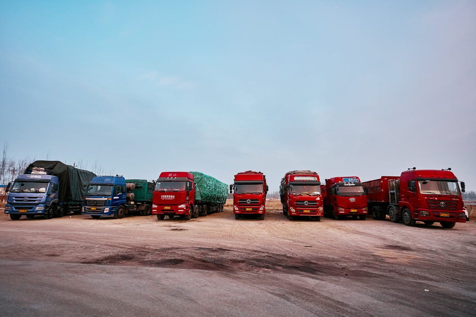 Row of seven large industrial trucks parked on a dirt lot. The trucks are a mix of colors including blue, green, and predominantly red. They appear to be transport vehicles for agricultural or industrial goods, with some carrying covered loads. The scene is set against a clear blue sky, suggesting an open rural or industrial area.