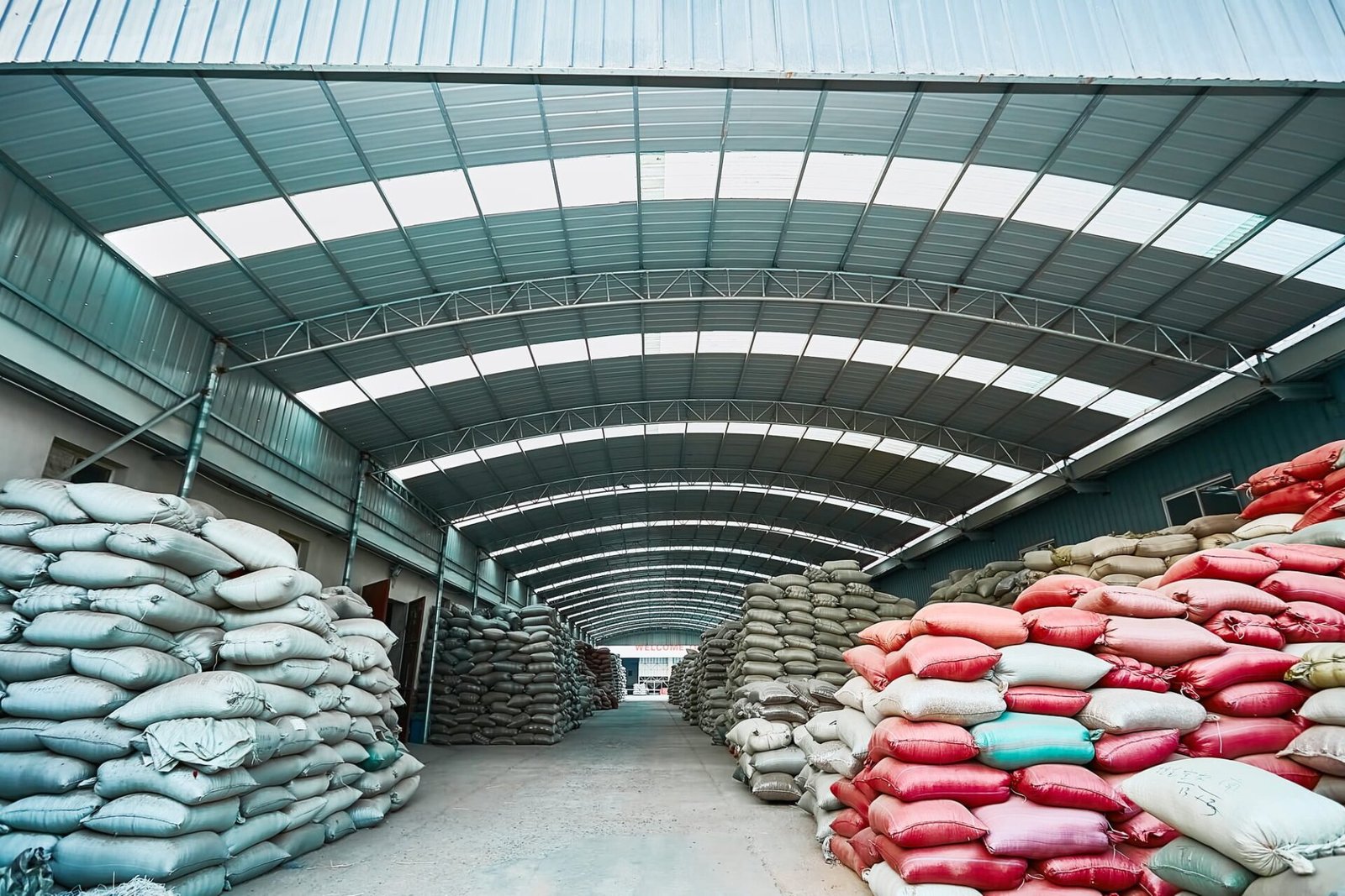 Interior view of a spacious, covered warehouse with an arched metal roof. Stacks of colorful large sacks, likely containing agricultural products, line both sides of a central aisle. The sacks are in various colors including white, gray, red, and blue. The warehouse structure provides protection from the elements while allowing for organized storage and easy access to the goods.