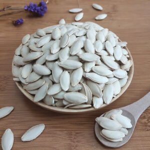 A ceramic plate filled with white, hull-on pumpkin seeds is displayed on a wooden surface. The seeds are oval-shaped with a slightly raised center, showing their natural, unprocessed state. A ceramic spoon holding a few seeds rests nearby, suggesting readiness for consumption. In the top left corner, a small sprig of purple lavender adds a delicate contrast to the pale seeds. Some seeds are scattered around the plate, emphasizing abundance. This image showcases the pure, natural appearance of pumpkin seeds in their shell, highlighting their potential as a nutritious snack or ingredient.