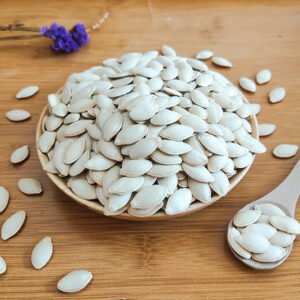 A wooden plate heaped with white, hull-less pumpkin seeds dominates the image. The seeds are flat and oval-shaped, with a subtle sheen. Some seeds spill onto the light wooden surface beneath. A wooden spoon filled with seeds rests nearby, emphasizing their ready-to-eat nature. In the top left corner, a small sprig of purple lavender adds a touch of color contrast. This image showcases the clean, pure appearance of hulled pumpkin seeds, suggesting their potential as a healthy snack or ingredient.