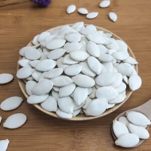 A ceramic plate overflows with pristine white pumpkin seeds on a wooden surface. The seeds are hull-on, showcasing their natural oval shape with slightly raised centers. Their uniform white color gives them a clean, fresh appearance. A ceramic spoon filled with seeds rests in the bottom right corner, suggesting readiness for consumption. A few seeds are scattered around the plate, emphasizing abundance. In the top left, a small sprig of purple lavender provides a subtle color contrast. This image highlights the pure, unprocessed nature of these pumpkin seeds, presenting them as a wholesome snack or ingredient.