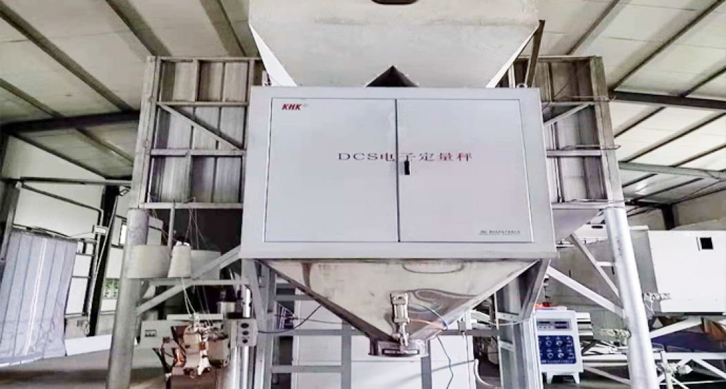 Industrial automated seed packaging system in operation. The image shows a large machine with multiple chutes feeding into bags. Workers are monitoring the process as seeds or grains are being automatically weighed and packaged into uniform bags. The system appears to be designed for high-volume, precise portioning of agricultural products.